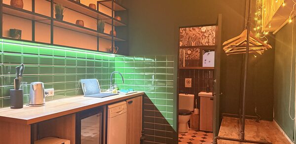 Лофт для вечеринок с коктейльным баром . Кухонное пространство с деревянной мебелью и барной стойкой, зелёная плитка на стенах, открытые полки с посудой и декором. Освещение желтоватого оттенка. Видна дверь в санузел, частично облицованный плиткой с узором. Напольное покрытие из тёмного дерева.