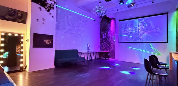 Лофт для вечеринок с коктейльным баром . Просторное помещение с деревянным полом, белыми стенами и декоративной подсветкой. Натуральный и диско-шар освещение. Мебель: диван, барные стулья и стол. На стене большой экран с проектором. Декор: картина, неоновая надпись и зеркало с лампами.