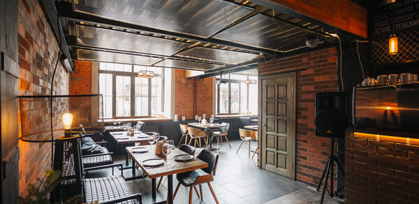 Двухуровневый лофт-бар. Просторное помещение с кирпичными стенами и металлическим потолком. Несколько деревянных столов с сервировкой, окруженные стульями и креслами. Панорамные окна с естественным освещением, подвесные светильники, темный плиточный пол.