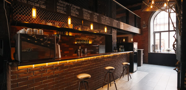 Двухуровневый лофт-бар. Барная зона с кирпичной барной стойкой, четырьмя высокими табуретами, подвесным освещением. На верхней полке стеклянные изделия и напитки. Стены из кирпича, большое арочное окно, меню написано на табличках над стойкой, тёмные потолок и пол.