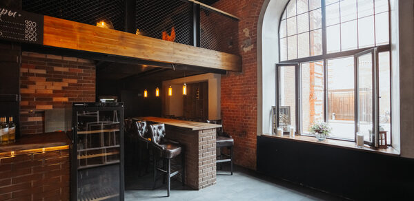 Двухуровневый лофт-бар. Промышленное пространство с кирпичными стенами, высоким потолком и большими арочными окнами. Барная стойка с кожаными стульями, подвесные светильники, металлические и деревянные элементы. На заднем фоне небольшая стойка и ценники. Кафельный пол серого цвета.