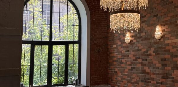 Geo loft (Гео Лофт). Кирпичный лофт с большими окнами. Арочное окно с черной рамой, кирпичная стена разных оттенков, два хрустальных светильника, кожаные кресла коричневого цвета, дневной свет, две декоративные свечи на подоконнике.
