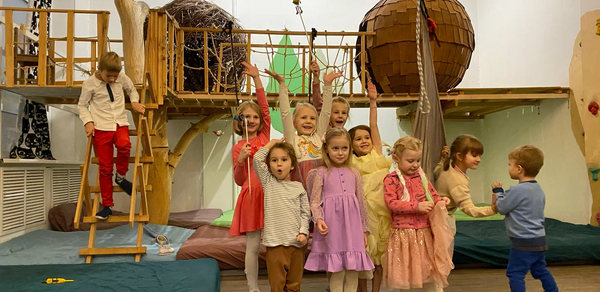 Children's eco-loft. Детская игровая комната с деревянными конструкциями, подвесными мостами, лестницей и сеткой. Светлые стены, деревянный пол. Группа детей в яркой одежде на переднем плане. Декоративные элементы в виде больших плетеных шаров.