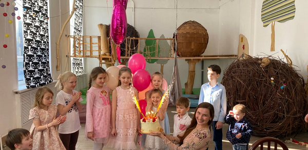 Children's eco-loft. Детская игровая комната с деревянными игровыми конструкциями и большой корзиной из веток. Светлые стены с рисунками деревьев, белые столы, цветные шары и занавески с узором. Освещение естественное, пространство оформлено для праздника.