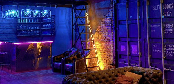 Контраст Loft. Диваны, стол. Декорация в виде дверей большого контейнера. Большая навесная люстра с несколькими лампочками. . Телевизор. Стулья. Свёрнутый экран на стене