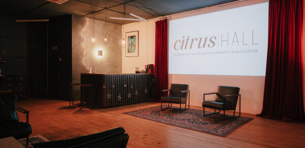 Citrus Hall (Цитрус Холл). Лофт Грейпфрут. Пространство с деревянным полом и тёмным потолком. Экран с надписью, два кресла на ковре, барная стойка с декоративной подсветкой. Красные шторы, подвесные светильники, картина на стене. Интерьер в тёмно-красных и чёрных оттенках.