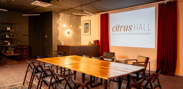 Citrus Hall (Цитрус Холл). Лофт Грейпфрут. Многофункциональное пространство с деревянным столом, складными стульями, проектором на белой стене с красными шторами. Слева барная стойка с подвесными светильниками, стеллаж и шкаф. Потолок темный, пол деревянный с ковром.