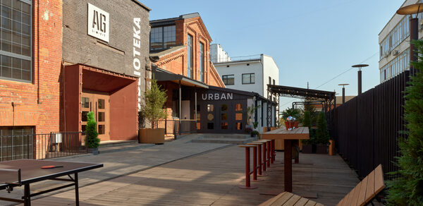 AG Loft. Urban (АГ Лофт. Урбан). Просторный лофт с раритетным автомобилем. Открытая терраса с деревянным настилом, столами и скамейками; кирпичное и серое здание с большими окнами; вывески "AG" и "URBAN"; стол для пинг-понга; растения в горшках; ясное небо, хорошее освещение.