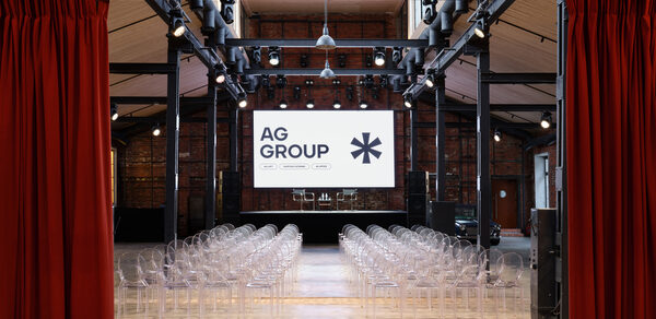 AG Loft. Urban (АГ Лофт. Урбан). Просторный лофт с раритетным автомобилем. Просторное помещение с высокой крышей и кирпичными стенами, разделанное металлическими балками. Центр оснащен большим экраном с надписью "AG Group". Установлены прозрачные пластиковые стулья, расположенные рядами. Освещение представлено подвесными светильниками. Между колоннами висят закрытые красные шторы.