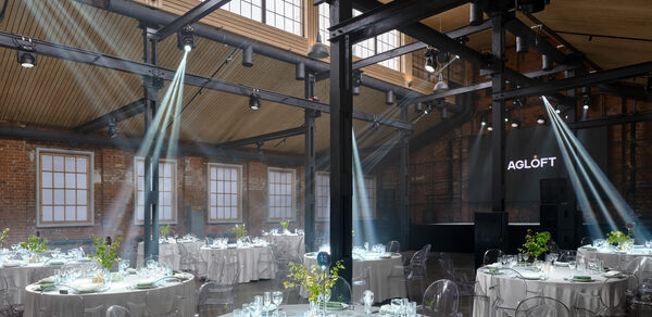 AG Loft. Urban (АГ Лофт. Урбан). Просторный лофт с раритетным автомобилем. Просторное помещение в стиле лофт с кирпичными стенами и высокой деревянной крышей. Несколько круглых столов с белыми скатертями и прозрачными стульями. Яркое освещение, световые лучи, окна под крышей. Надпись "AGLOFT" на черной стене.