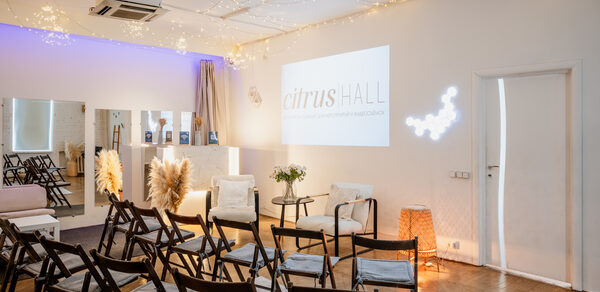 Citrus Hall (Цитрус Холл). Зал Свити. Светлое помещение с деревянными складными стульями, расставленными в рядах. Белые стены с проектором. На потолке гирлянды светодиодных огней. Зона с зеркалами, декоративные элементы из сухоцветов, плетеная лампа.