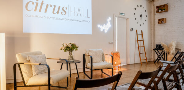 Citrus Hall (Цитрус Холл). Зал Свити. Просторное помещение с деревянным полом, белыми кирпичными стенами и проектором. Есть стулья, кресла и круглый стол с вазой и цветами. Декоративные элементы: лестница, настенные часы, световые гирлянды и крупный текст проекта.