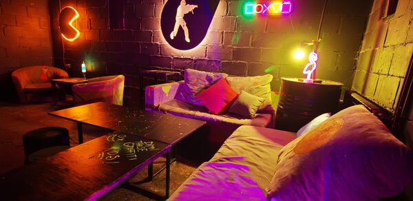 Лофт Геймеры. Крупный план диванов, между ними стоит бочка. Рядом расположен стол, вдалеке стоят кресла и ещё один стол. На стене неоновые лампы и игровые логотипы