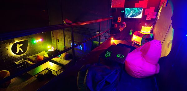 Лофт Геймеры. Второй этаж лофта. К стене прикреплены небольшие картины с телевизором. На полу стоит небольшой столик с пуфами, в углу маленькая тумбочка с лампой. Также видно убранство первого этажа