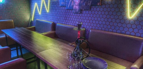 Лофт Соты. Пространство с длинным столом и кожаными диванами вдоль стены. На столе кальян, тарелки и бокалы. Стена отделана шестигранной плиткой, украшена неоновыми линиями и картинами. Освещение синими и жёлтыми тонами.