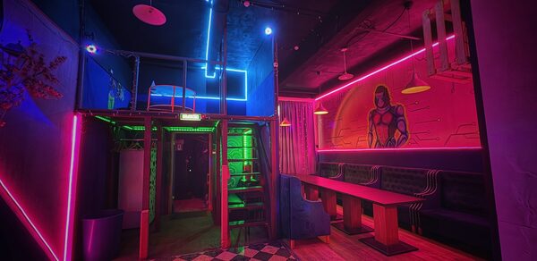 Лофт в стиле Диско. Лофт-пространство с черно-белым клетчатым полом. Неоновая подсветка розовых и синих цветов. Лестница на второй уровень, на стене стена с граффити героя. Диваны и длинный стол. Потолочные светильники. Тёмный потолок.