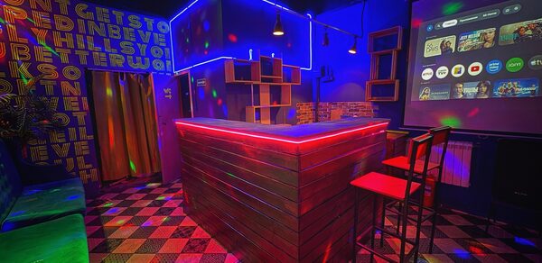 Лофт в стиле Диско. Караоке-зал с барной стойкой, оформленной деревом и подсветкой. Зал включает экран с интерфейсом приложений, разноцветные стулья и диван. Стены с яркими надписями, плиточный пол геометрического узора, разноцветное освещение.