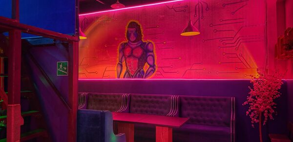 Лофт в стиле Диско. Лофт-пространство с ярким неоновым освещением, стеной, украшенной граффити с изображением человека. Темно-синие диваны вдоль стены, деревянные столы, красные и синие акценты, подвесные светильники, лестница, указатель выхода, искусственное растение.