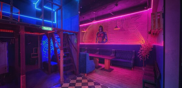 Лофт в стиле Диско. Клубное пространство с неоновым освещением в синих и розовых тонах. Деревянные лестницы ведут на второй этаж. Черно-белая плитка на полу, мягкие диваны вдоль стены. Настенные граффити, висят светильники, знак "Выход".