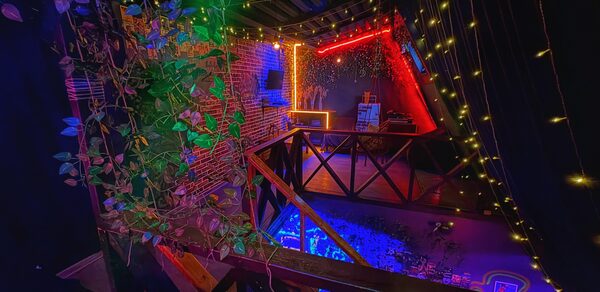 Лофт-бар. Крытое пространство с кирпичными стенами, балконом с деревянными перилами и растениями. Яркое разноцветное освещение, гирлянды на потолке. Обстановка с барной стойкой, стульями, квадратным столиком и черными занавесками.