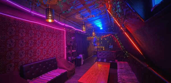 Лофт-бар. Клубное пространство с затенённой мебелью, включающей кожаные диваны и деревянный стол. Яркое неоновое освещение в синей и фиолетовой гамме. Декор из искусственных растений на потолке. На стенах тёмные панели и узорчатый ковёр.