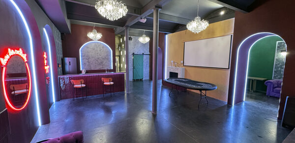 Белый лофт с яркой мебелью. Многофункциональное пространство с барной стойкой и высокими стульями, проектором на стене, стилизованными арками с неоновой подсветкой. Потолочные люстры, пол из темного материала, стены в теплых тонах.