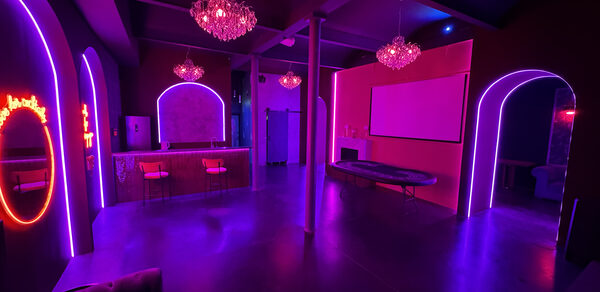 Белый лофт с яркой мебелью. Пространство с неоновым освещением, лофт-стиль, фиолетовые и розовые цветовые акценты. Арочные проемы, зеркала, барная стойка с двумя высокими стульями. Потолочные люстры, белый экран на стене, круглый стол. Полы глянцевые, металлические колонны.