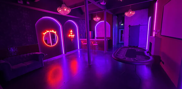 Белый лофт с яркой мебелью. Клубное пространство с неоновым освещением фиолетового и розового цветов, настенные неоновые вывески, диван, барная стойка с высокими стульями, стол для игры в рулетку, декоративные люстры, затемненный интерьер, стены с темными обоями, потолок с балками.