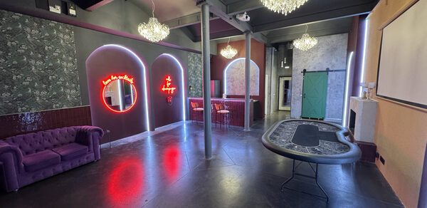 Белый лофт с яркой мебелью. Лофт-пространство с темным потолком и полом. Фиолетовый диван, покерный стол, барная стойка со стульями. Неоновые надписи на стенах, цветочные обои, арочные ниши, зеркала. Люстры с хрустальными элементами. Зеленая дверь-купе.