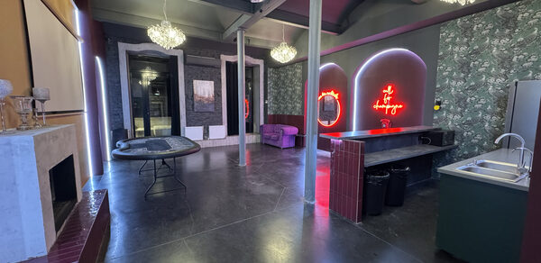 Белый лофт с яркой мебелью. Многофункциональное помещение с высокими потолками, серыми колоннами и темным полом. Есть камин, круглый стол, барная стойка с раковиной, фиолетовый диван. На стенах обои с ботаническим узором, светильники, неоновая вывеска.