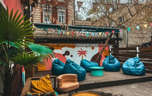 Лофт Surf (Серф). Тропический лофт с большой верандой.. Открытая терраса с деревянным настилом, украшенная гирляндами из треугольных флажков. Мебель: мягкие кресла и яркие пуфы, круглый столик. На фоне разноцветная стена с пальмовым рисунком. Расставлены растения в горшках.