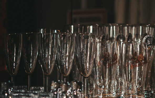 Бар под закрытие со сценой . Стеклянные бокалы для шампанского и пивные кружки выстроены в ряд. Прозрачное стекло, часть бокалов с логотипами. Тёмный фон, тёплое освещение.
