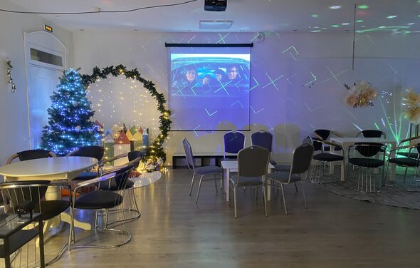 Лофт для ярких душевных вечеринок. Просторная комната с круглыми столами и стульями, новогодней елкой и светящимися декорациями слева. На стене проецируется изображение. Светлые стены, деревянный пол, разноцветное декоративное освещение.
