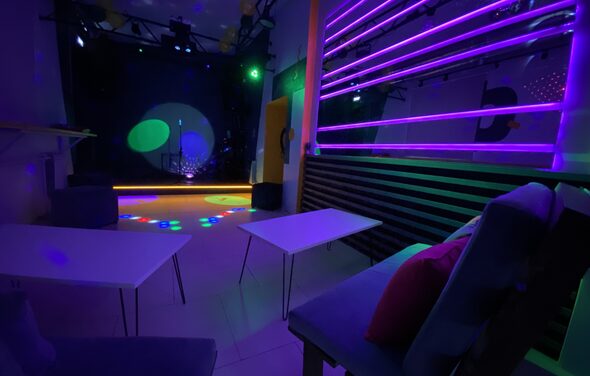 Музыкальный лофт на Тульской. Клубное помещение с фиолетовой и зеленой подсветкой, столами и мягкими креслами, черным стеновым покрытием, сценой с микрофоном и световыми эффектами на полу. На стене вертикальные неоновые полосы, на диванах разноцветные подушки.