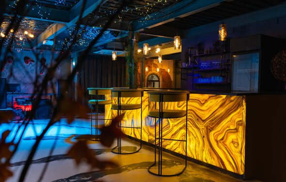 Стилизованный двухэтажный лофт. Современное пространство с барной стойкой из подсвеченного камня, высокими стульями, подвесными светильниками. На потолке гирлянды, деревянные элементы, различные зоны освещения. Интерьер в теплых и холодных тонах, видна декоративная растительность.