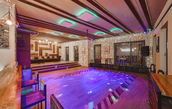 Loft Lodge 6. Просторное помещение с деревянным паркетом и ступеньками на возвышение. Деревянные балки на потолке с подсветкой. Барная стойка с высокими стульями. Неоновое и точечное освещение. Кожаные диваны, стулья, столики и аудиооборудование. Декоративные элементы на стенах.