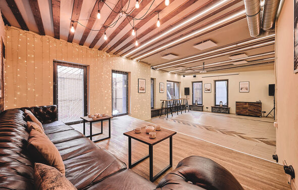 Loft Lodge 6. Просторная комната с деревянным полом и полосатым потолком. Тёмно-коричневый кожаный диван с подушками, два столика. Стены декорированы гирляндами и картинами. Оборудованная барная стойка и аудиосистема. Освещение из подвесных ламп. Трубы на потолке.