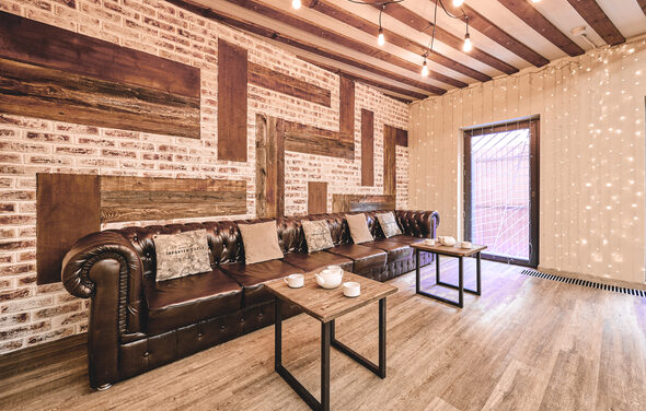Loft Lodge 6. Комната с кирпичными стенами и деревянными вставками, деревянный пол. Большой кожаный диван коричневого цвета с подушками, перед ним два столика с посудой. Освещение — лампочки на проводах, гирлянда на стене. Цвета тёплые, интерьер в отличном состоянии.