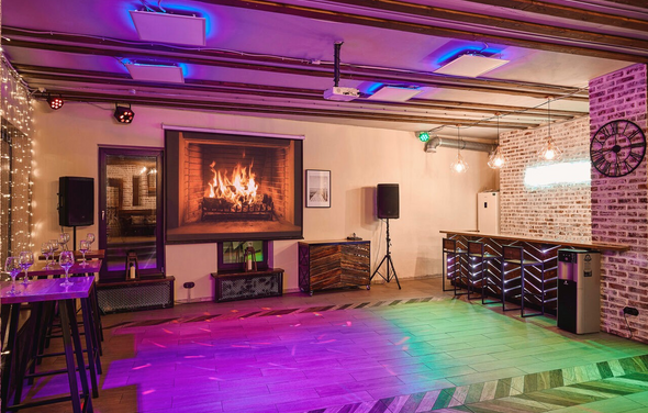 Loft Lodge 6. Это просторное помещение с деревянным полом и декором из кирпича. На стене изображение камина. Имеется барная стойка, столы и стулья. Освещение состоит из ламп и цветных прожекторов. Присутствуют аудиоколонки. На стене часы. Состояние ухоженное.