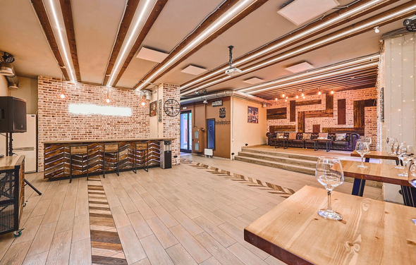 Loft Lodge 6. Это интерьер кафе или ресторана с кирпичными стенами. В центре деревянная барная стойка с высокими стульями. Потолок украшен деревянными балками и лампами. На полу плитка с узором. Присутствуют деревянные столы с бокалами. Общее освещение теплое.