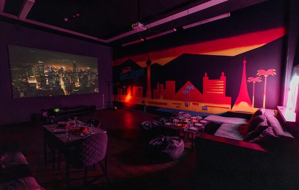 Зал Лас-вегас. Лофт-зал в американском стиле . Кинотеатр с проектором и большим экраном, декоративная стена с изображением городского пейзажа и неоновых огней, темная мебель: столы и диваны, расставленные по пространству, мягкие пуфы, приглушенное освещение, темные тона с яркими акцентами.