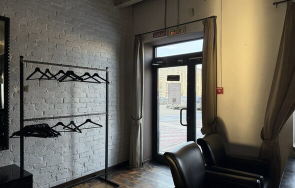 Лофт Повод Есть. Гардеробная зона с белой кирпичной стеной и металлической вешалкой с черными плечиками. Справа стеклянная дверь с вывесками «вход» и «выход», занавески на окнах. Деревянный пол, кожаные кресла темного цвета. Светильников не видно.