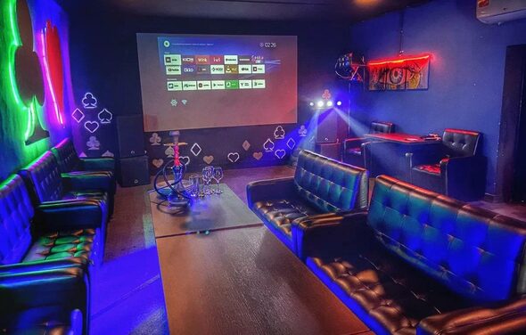 Rave(Рейв). Тёмное помещение с синими стенами, диваны с кожаной обивкой, проектор с интерфейсом на экране, деревянный стол, кальян. Неоновая подсветка, на стенах символы игральных карт. Потолочный и настенный светильники. Ковёр на полу.