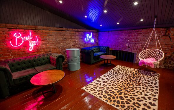 Двухэтажный лофт с отдельным входом. Лофт-пространство с кирпичными стенами. Две кожаные диваны, два круглых деревянных стола, подвесное кресло, ковер с леопардовым узором. Неоновая надпись на стене, металлическая бочка. Потолочное точечное освещение, темное деревянное покрытие пола и потолка.