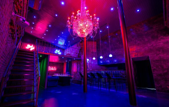 Двухэтажный лофт с отдельным входом. Двухуровневое помещение с кирпичными стенами и металлической лестницей. Потолок с точечным и неоновым освещением, люстра. Барная стойка с высокими стульями. На стенах неоновые надписи. Темно-красные и синие оттенки.