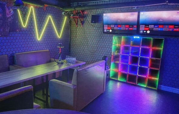 Bee-lounge(Пчела-лаундж). Караоке-зал с мягкими диванами и длинным деревянным столом. На стенах неоновая подсветка и светодиодные панели. Два телевизора на стене. Темный пол и сетчатые обои. Стеклянные стаканы и кальян на столе.