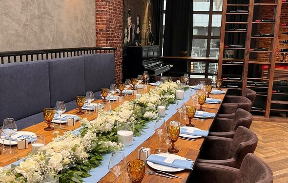 Площадка для мероприятий в стиле лофт. Длинный деревянный стол с цветочной композицией и сервировкой для мероприятия; коричневые кресла, кирпичная стена, синяя обивка дивана, пианино у окна, большая полка, стеклянные бокалы, свечи, голубые салфетки.