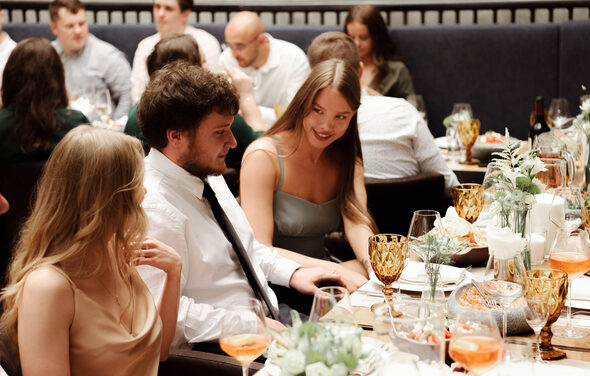 Площадка для мероприятий в стиле лофт. Большое количество людей за накрытым столом, сервировка с бокалами, тарелками и декоративными растениями. За столом темные стулья, на заднем плане видна мебель с высокими спинками. Освещение теплое, цвета нейтральные.