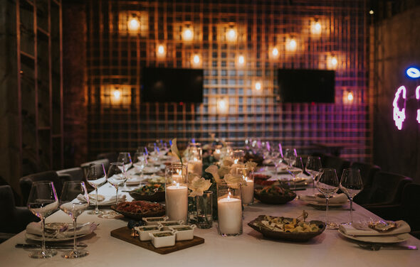 Площадка для мероприятий в стиле лофт. Длинный стол в зале с решетчатыми стенами, сервирован белой скатертью, украшен свечами и посудой. Потолочные светильники создают мягкое освещение. В центре стола блюда с едой. На стене два экрана и неоновая надпись.