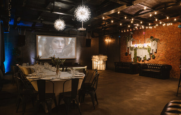 Nord Loft(Норд Лофт). Лофт-пространство с кирпичной стеной и индустриальным потолком. Стол с белой скатертью и стульями, кожаные диваны. Подвесные светильники и гирлянды. Проектор и экран. Неоновая вывеска и декоративные элементы на стене. Тёплый свет.